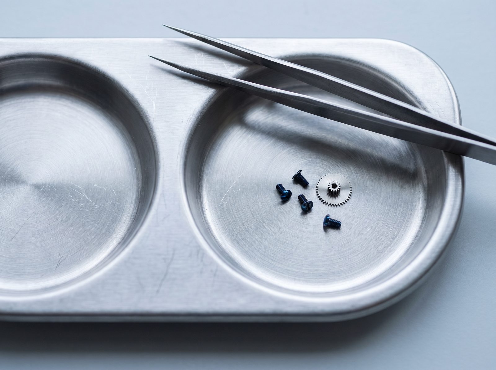 Watchmaker's tweezers over a steel parts tray holding blued screws and a small wheel.